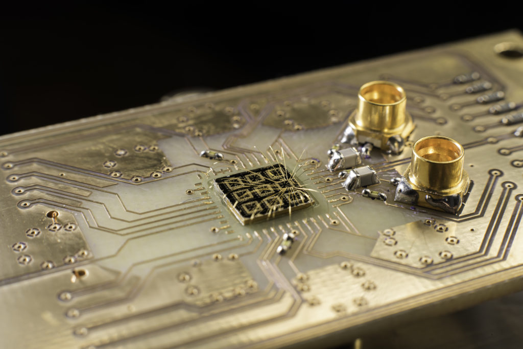 Thin aluminum wires connect the surface of a quantum processor semiconductor chip to pads on a circuit board. The researchers fabricate the device by patterning and depositing metal gates on a chip. The metal gates are designed to trap individual electrons in the semiconductor. The researchers send electrical signals to the device via the aluminum wires, changing the voltage on the metal gates to control the electrons. They also receive electrical signals from the device to help monitor the electrons’ behavior. (University of Rochester photo / J. Adam Fenster) Thin aluminum wires connect the surface of a quantum processor semiconductor chip to pads on a circuit board. The researchers fabricate the device by patterning and depositing metal gates on a chip. The metal gates are designed to trap individual electrons in the semiconductor. The researchers send electrical signals to the device via the aluminum wires, changing the voltage on the metal gates to control the electrons. They also receive electrical signals from the device to help monitor the electrons’ behavior. (University of Rochester photo / J. Adam Fenster)